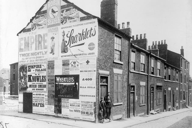 This view shows a row of terraced housing on the north-east side of Nile Street, taken during improvements to the North Street area. A man and boy stand outside the doorway of the end house. The gable end of the block is covered with advertising, including a poster for the Leeds Hospital Bank Holiday Gala and Sports at Roundhay Park, on Monday, August 6 and Tuesday, August 7, 1900. Back Nile Street leads off Nile Street in the foreground then continues around the back of the houses, with Brunswick Row leading off this on the far left.