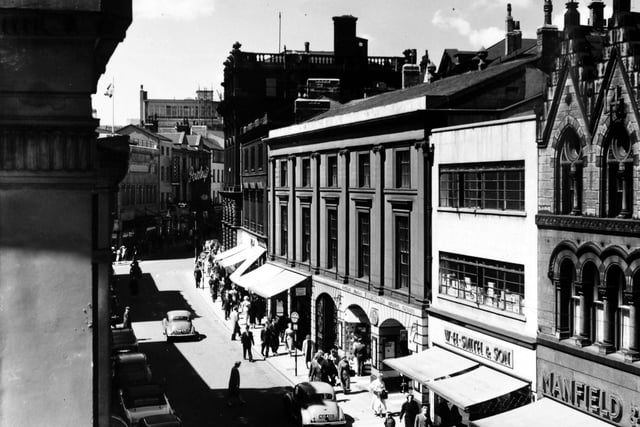 A view of Commercial Street from an upper storey window in may 1957. Various shops and businesses are visible, including, at the right edge, Manfield & Sons Ltd., shoe retailers at number 14 Commercial Street. Moving left, the newsagents business of W.H. Smith & Son Ltd occupies number 16 and the ground floor of number 18 (with the arched entrance and windows). Leeds Library has occupied the upper storeys since 1808. The view is towards the junction with Albion Street.