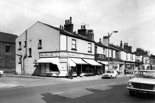 A view looking down Dewsbury Road towards Hunslet Hall Road. Shops include Harry Norris family butcher at number 164a with fenton dispensing chemist at number 162. A pram stands outside the butchers while a man, woman and child walk towards Roxburgh Road. Cars are parked at the curb while people walk past the shops. In the distance an advertisement for Mackesons Ale can be seen. Pictured in August 1964.