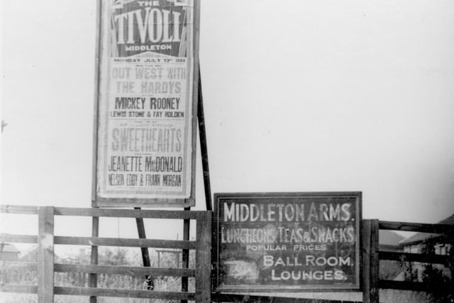 A low timber fence on Middleton Park Avenue. There is a painted wooden sign offering directions to the Middleton Arms, and beside this a free-standing hoarding promoting films at the Tivoli. Pictured in July 1939.