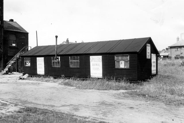 A one-storey wooden construction in Dunderdale Yard in March 1965. Signs for Spiller's Animal Foods are visible. The building also has a venting chimney through one of the windows. On the left edge, the former Crown Chemical Works premises can be seen.