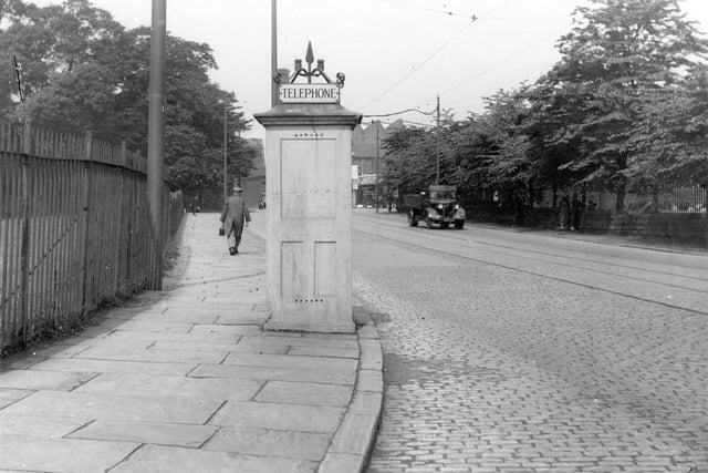 Telephone kiosk at the junction of Stanningley Road and Henconner Lane, looking towards Stanningley Road in June 1936. To right of picture are the grounds of Good Shepherd School, the annexe of Christ the King R. C. School, bounded by a wall with iron railings and trees behind.  A man is walking along the pavement.