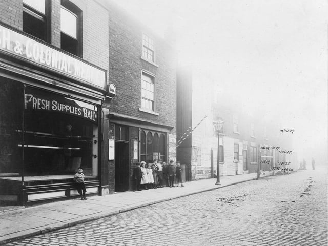 A group of children look at the camera on Waterloo Road. On the left is a butchers shop. In the centre, barbers poles and wall sign 'Junction Shaving Saloon'.