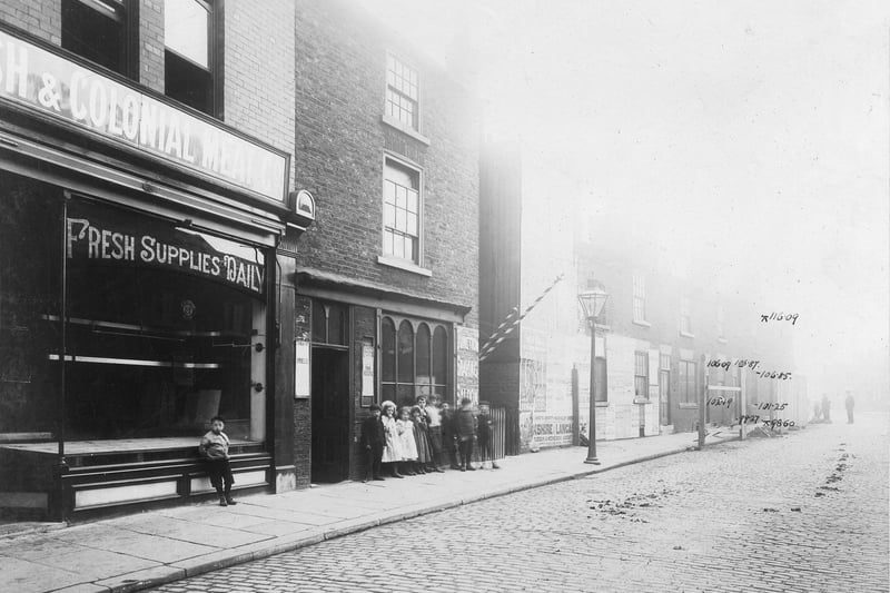A group of children look at the camera on Waterloo Road. On the left is a butchers shop. In the centre, barbers poles and wall sign 'Junction Shaving Saloon'.
