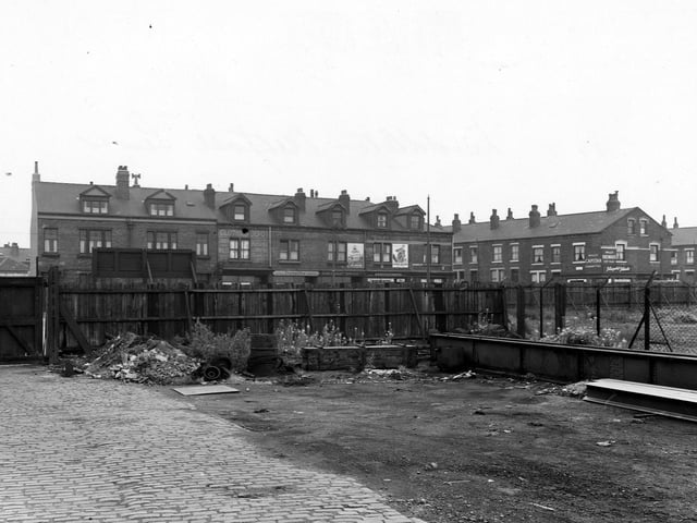 Looking across the part cobbled yard of Coghlan Forge and Rolling Mills Ltd, to a wooden fence with large wooden gate, and chicken wire fence to the right. Beyond the fence is a row of shops on Low Road. These are the Clothing Depot, Dorothy, ladies hairdresser, Don's Valet Service, dry cleaners, A.E. Forster and Son, grocers and Jas. Wm. Walton, cycle agent. The street on the right is Iveridge Street.