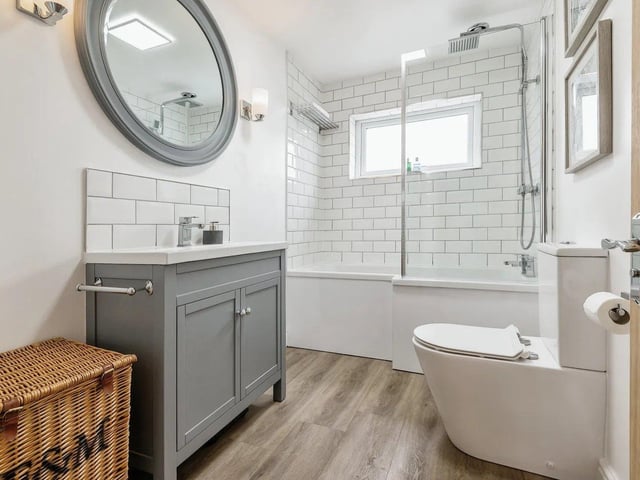 The modern white suite comprises a P-shaped bath with glazed shower screen, vanity wash basin, low level WC, tiled surrounds and chrome towel rail.
