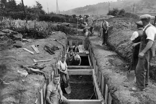 An excavation for a sewer near Meanwood Road. Several workmen are standing in and around a large trench. Woodhouse Ridge can be seen in the background as can a factory chimney. Pictured in July 1937.