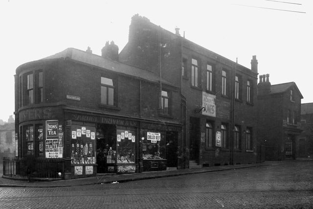 Photo taken from Domestic Street of Shafton Lane in January 1929. Parade of shops; starting from the left - no 1; Samuel Driver and Co - a grocers shop. Then 1A Shafton Lane - British and Argentine Meat Co butchers. Then a taller building which is Holbeck Pitt Conservative Club. A big poster on the outside of the building says to vote for Albert E Jones in the northern elections. To the right is 5A Shafton Lane - a greengrocers run by Jack Smith. Tramlines are visible on the wet, cobbled road.