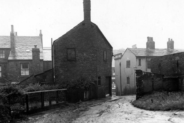 The Gang located at the back of Town Street. A muddy road is visible leading up to to the foreground from Town Street. The backs of houses are visible on the left with a garden and bushes etc. The premises of 'Isaac Stephenson Ltd. Butchers' can be seen through the gap. Pictured in August 1952.
