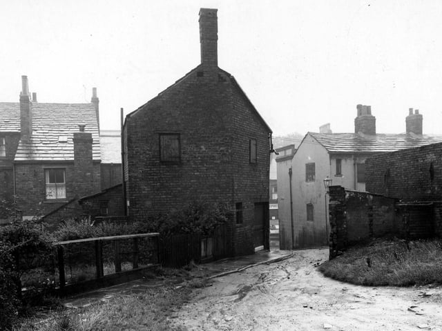 The Gang located at the back of Town Street. A muddy road is visible leading up to to the foreground from Town Street. The backs of houses are visible on the left with a garden and bushes etc. The premises of 'Isaac Stephenson Ltd. Butchers' can be seen through the gap. Pictured in August 1952.