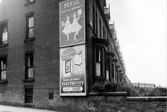 A view looking south at the gable end of number 171 Cardigan Road from Burley Lodge Road in September 1953. The houses are terraced. Hoardings advertising Persil and The Yorkshire Electricity Board are to the right of a small metal plate covering a coal delivery hole.
