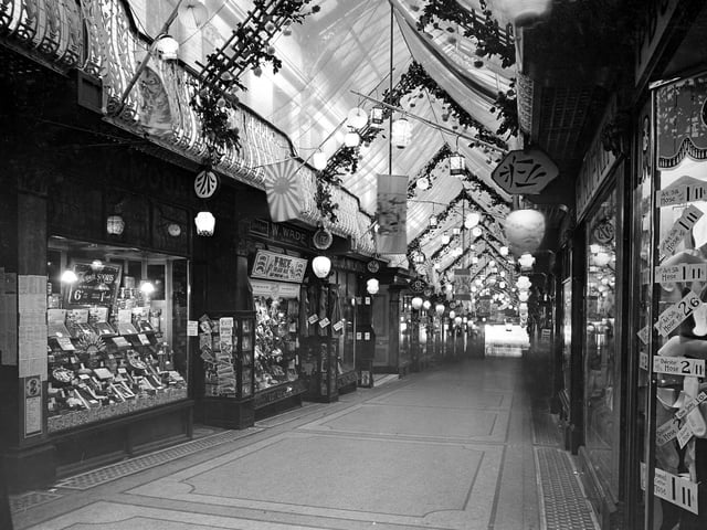 The interior of the Queen's Arcade decorated in a Japanese style for Civic Week. Lanterns are hanging from the balcony where a fabric canopy has been constructed. Trellis arches span the arcade decorated with chrysanthemums which are one of the national flowers of Japan. On the left are W.Mooney and Sons., confectioners, and W.Wade, outfitters and hatters. On the right, Leah Rifkin's drapers is visible. The view looks down from Lands Lane towards Briggate.