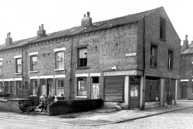 The corner of Ascot Terrace and Cross Ascot Street in October 1966. On the left is the rear garden and entrances of through terraces fronting onto Ascot Avenue. Four children play in the run-down garden of derelict number 68. The shop at number 70 is also derelict with boarded up windows and door downstairs and missing windows in the first and second floor and crumbling brickwork. On the wall above the shop window is is a faded painted advertisement for Capstan Cigarettes. A woman walks along Cross Ascot Street on the right of the building.