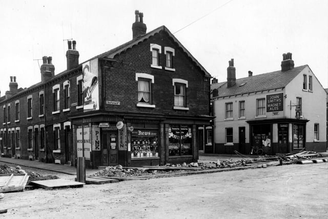 Shops along New Pepper Road in August 1956 showing where a trench has been excavated for sewerage. To the right can be seen end of Rocheford Terrace, with off licence at end. Advert for John Smith's Magnet Ales on wall. Greengrocers and newsagent on opposite corner.