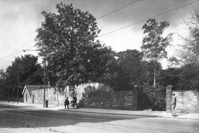 Canal Gardens on Princes Avenue in July 1930. Stone wall runs down the side of a pavement alongside Canal Gardens. Two men, one with a bike walk down the pavement with another man in period dress coming down the other way. There is a water pressure meter on the wall and a gateway leading into Canal Gardens. Two sets of tramlines run down Princes Avenue.