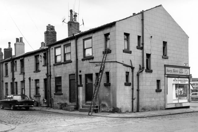 Back-to-back terraced houses at the rear of shops on Dewsbury Road in August 1964. A car, reg: 916 HUB is parked on left watched by a dog stood on the step of number 23, two ladders are propped up against the side of number 27. On the right is Roxburgh Road leading to Dewsbury Road. Visible on the right is number 164a Dewsbury Road, Harry Norris family Butcher, with hygienically refrigerated counters.