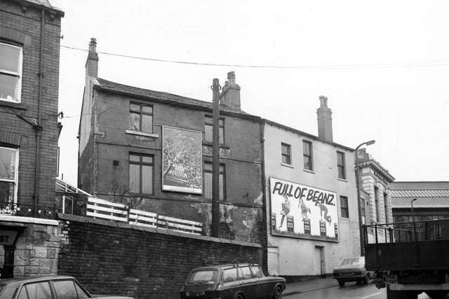 Crab Lane showing the rear of buildings numbered 15-17 Stocks Hill. Advertisements for Winston cigarettes and Heinz baked beans are on the walls. To the right of the picture is the junction with Stocks Hill, with the Midland Bank on the corner; part of Armley Library can be seen on the far right. Cars and a truck are on the road. Pictured in September 1982.