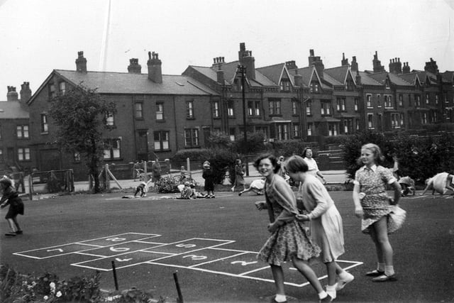 A group of three schoolgirls in the playground of Burton House, playing near an area marked out for games of Hopscotch. In the background, a terraced street of red brick back-to-back houses situated off Burton Avenue is seen. This is the even-numbered side of Fairford Avenue.