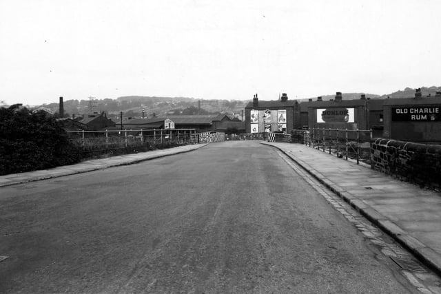 Armley Ridge Road in September 1949 looking north to Amen Corner. The left-hand pavement is bordered by bushes and railings leading to stone walls painted in black and white check at Amen Corner. The right-hand pavement has stone walls and railings to the side. To the right beyond the road, are the backs of three rows of houses, with advertising posters on them. The middle one is for Dunlop, the one on the right is for Old Charlie Rum. On the left, beyond the road, can be seen Hirst and Thackway Ltd., weather coat manufacturers, the Leeds Briquette Works, and the London, Midland and Scottish Railway Co. Goods