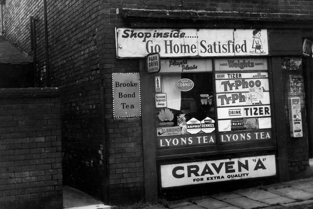 The grocers at 201 Jack Lane pictured in August 1963. Advertised above the shop on posters and signs are products including Burwicks baking powder and Robinsons Lemon Barley. Above the shop window is the slogan 'shop inside and go home satisfied' while below signs promote Park Drive cigarettes, Brooke Bond tea, Players Weights cigarettes, Senior Service cigarettes, banana's, Typhoo tea and Tizer and Ogden's Cobnut, St. Bruno's and Walnut tobacco. Aspro and Rennies tablets are promoted alongside Lyons tea and Craven 'A' cigarettes. A bubble gum machine stands by the shop door on the right while on the far left of the building there is a yard, originally built to house the shared outside toilets.