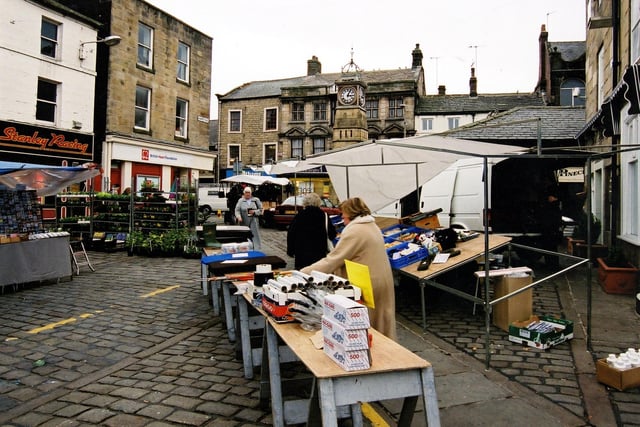 A view of stalls in Otley Market Place looking in the direction of Kirkgate. In the middle distance, centre is the Buttercross and the Jubilee Clock erected in 1888 to mark the Golden Jubilee of Queen Victoria. The clock tower was designed by Alfred Marshall and built by Mr W. Maston. It has four dials measuring three feet in diameter. The total cost of the clock and tower was £180.
