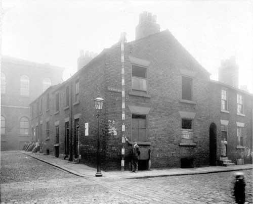 The junction of Lydia Street with St John's Square of derelict 2 storey terraced houses with cellars. Man stands with measuring pole to Gable End of house. Pictured in October 1910.