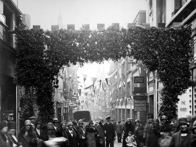 Trinity Street during Civic Week can be seen. Entered through an evergreen arch, the street is decked with bunting and busy with people. During Civic Week people were given the chance to look behind the scenes at businesses and educational establishments as well as Municipal departments and services including Whitehall Road Generating Station, Knostrop Sewage Disposal Works and Park Street Fire Station.