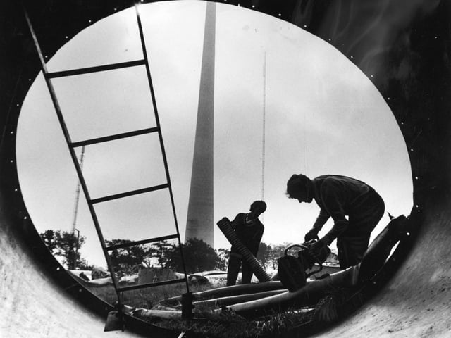 The new TV mast at Emley Moor, near Huddersfield, soars up through low cloud in September 1970 as John Orton and Derek Miller, framed in the broken section of the old mast start work on cutting up copper and aluminium cable. Work on clearing the wreckage of the old mast, which was brought down by snow and ice 18 months ago was delayed while the committee of inquiry completed investigations.