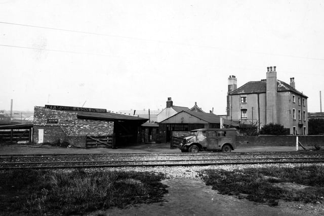 The premises of W.F. McQuire Ltd. (Road Transport Contractors) pictured in August 1947. In the distance there are factories with a house to the right, in front of them. There are two garages beside each other, and there is a Bannister Bros. Motor Body Builders van parked in front.