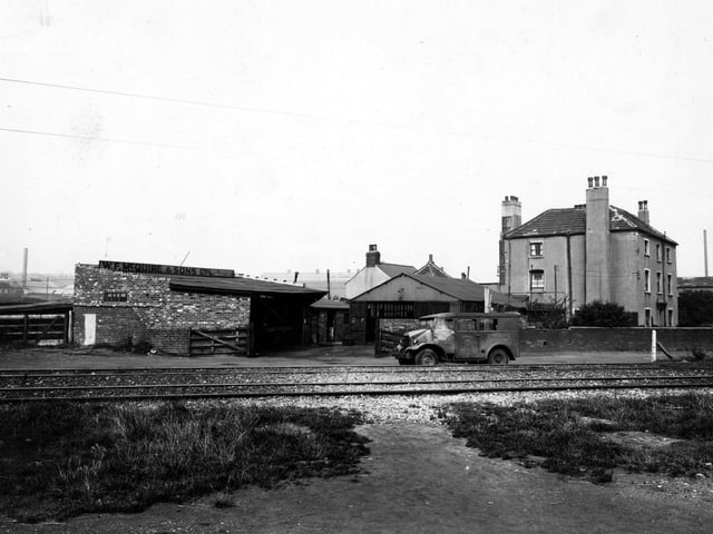 The premises of W.F. McQuire Ltd. (Road Transport Contractors) pictured in August 1947. In the distance there are factories with a house to the right, in front of them. There are two garages beside each other, and there is a Bannister Bros. Motor Body Builders van parked in front.