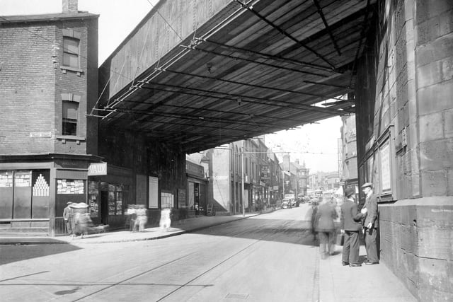 A view looking up Kirkgate from junction of Wharf Street. two men chat underneath railway bridge. On other side of road on corner of Wharf Street is L.Vaux wholesaler. Train timetables and details of excursions are posted on wall under bridge. Photograph shows a busy street with cars and a tram. Signs for Walkers drapers and William Brotherton and son can be seen.