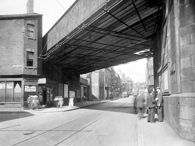 A view looking up Kirkgate from junction of Wharf Street. two men chat underneath railway bridge. On other side of road on corner of Wharf Street is L.Vaux wholesaler. Train timetables and details of excursions are posted on wall under bridge. Photograph shows a busy street with cars and a tram. Signs for Walkers drapers and William Brotherton and son can be seen.