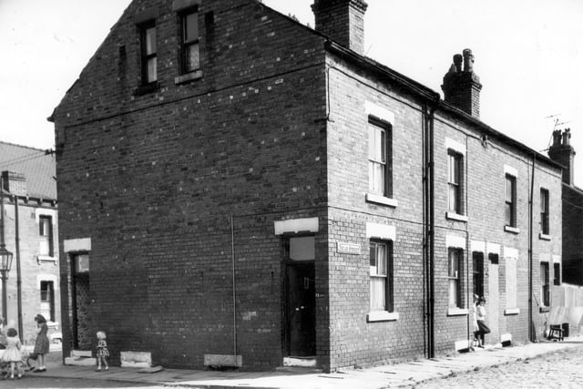The corner of Selborne Street on the left and Vicar Street on the right in August 1964. The two houses on the gable end of the property are numbers 19 and 21, both front doors are open and girls in summer dresses stand by a pram on the corner on the left. On the right two people stand in the doorway of number 10 Vicar Street while the door and window of number 8 on the right has been bricked up. Further down the street washing is hung out.