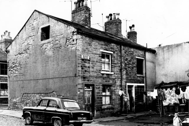 On the left of the image is School Place where a car, reg: ROH 346 is parked at the corner with Mann's Square on the right. Clothes hang on lines stretched across the square while number 3 in the corner appears empty and run down with a missing first floor window. On the far right behind the clothes lines a row of shared outside toilets.