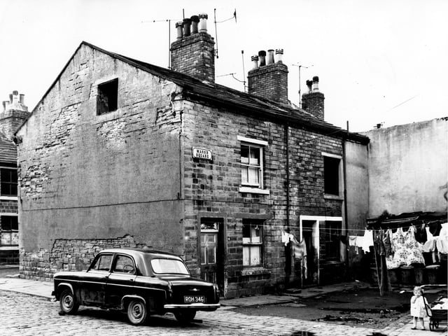 On the left of the image is School Place where a car, reg: ROH 346 is parked at the corner with Mann's Square on the right. Clothes hang on lines stretched across the square while number 3 in the corner appears empty and run down with a missing first floor window. On the far right behind the clothes lines a row of shared outside toilets.