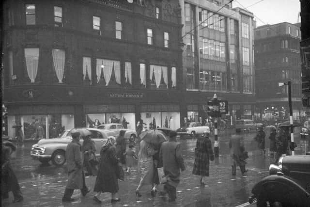 People crossing the road wearing rain coats and using umbrellas. Matthias Robinson department store in the background around 1960-1969.