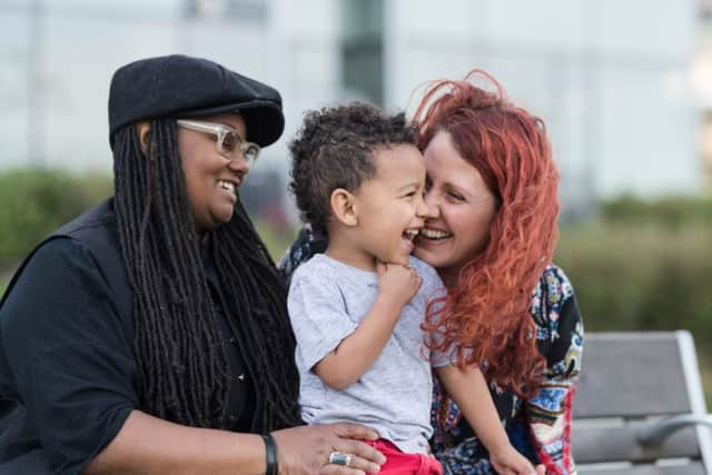 A homosexual biracial couple sit on a park bench with their young son. He is sitting on their laps and they're snuggling and talking with him. One mom is African-American and the other is Caucasian.